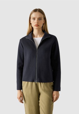 Frau mit glatten blonden Haaren, die eine marineblaue Jacke mit Reißverschluss über einem weißen Oberteil und beige Hosen trägt, vor einem schlichten Hintergrund stehend.