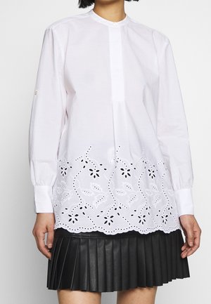 Vrouw die een witte blouse met lange mouwen en bloemrijke cut-out borduursel draagt, gecombineerd met een zwarte geplooide rok.