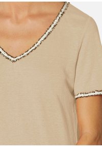 Beige T-shirt met een V-hals, voorzien van een gestructureerde rand in wit en bruin langs de halslijn en mouwen. Zacht, lichtgewicht stof.