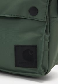 Grøn stofrygsæk med en glat overflade, der har et sort logo-patch og en knaplukning.