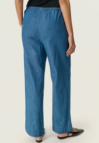 Denimbyxor med vida ben och elastisk midja, i en blå nyans med en slät textur, visade från baksidan med en svart sko.