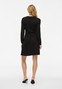 Rochie neagră cu mânecă lungă, tăietură dreaptă, fustă până la genunchi și textură moale. Purtați cu pantofi cu toc înalt negri și un decolteu simplu.