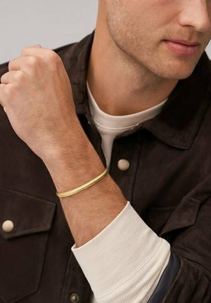 Gold-tone metal bracelet worn on a wrist, featuring a sleek, rounded design. The background shows a dark jacket and white shirt.