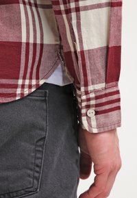 Gros plan sur le bras d'une personne portant une chemise à carreaux rouges et blancs avec le poignet boutonné, reposant à côté d'une poche de pantalon gris foncé.