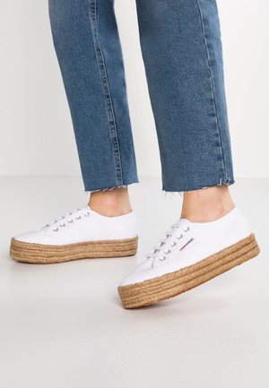Weiße Schnür-Plateausneaker mit geflochtenen Seilsohlen, getragen zu blauen, gekürzten Jeans auf hellem Boden.