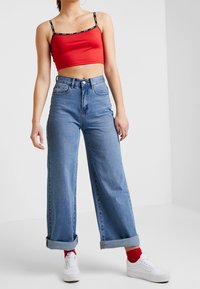 Débardeur court rouge avec bretelles noires, associé à un jean bleu clair taille haute à jambes larges, ourlets roulés, et baskets blanches.