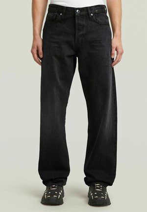 Personne portant un jean noir ample et des baskets noires et blanches, debout devant un fond clair uni.