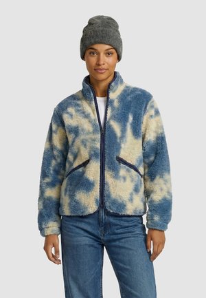 Jeune personne portant une veste zippée pelucheuse bleue et beige, un jean bleu et un bonnet gris en tricot, debout devant un fond uni.