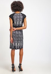 Vestido de estampado en blanco y negro con mangas tipo capucha, silueta ajustada y dobladillo en forma de ondas. Llevado con zapatos de tacón negros.