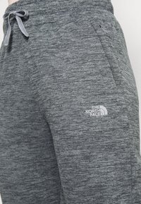 Grå träningsbyxor med en texturerad tyg, elastisk midja, dragband, sidofickor och en vit logotyp fr ån The North Face.