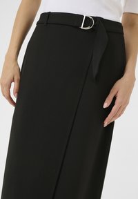 Jupe noire en tissu lisse, dotée d'une ceinture avec une boucle en argent et de détails de noeud sur le côté. Coupe ajustée avec un design longueur genou.