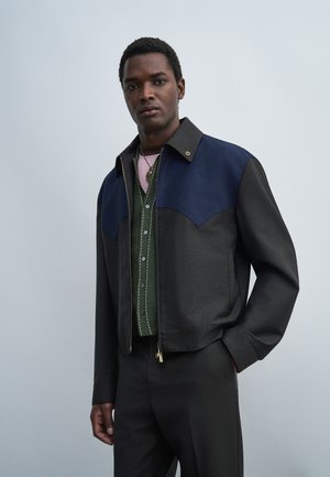 Mand iført en mørk jakke med mørkeblå skulderdel, grøn knappecardigan og pink skjorte, står med den ene hånd i lommen mod en ensfarvet baggrund.