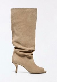 Bež semiš škornji srednje višine do meče z odprtim prstom in tankim petkim petkom na enostavni svetli podlagi.