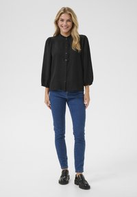 Schwarze Langarmbluse mit strukturiertem Stoff, Knopfleiste und Rüschensaum. Kombiniert mit blauen Skinny Jeans und schwarzen Schuhen.