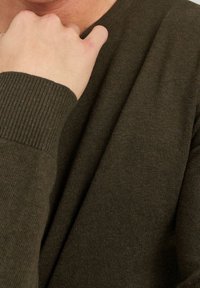 Maglione verde oliva con polsini a coste e texture liscia, che copre parzialmente una mano appoggiata vicino al mento. Design semplice e classico.