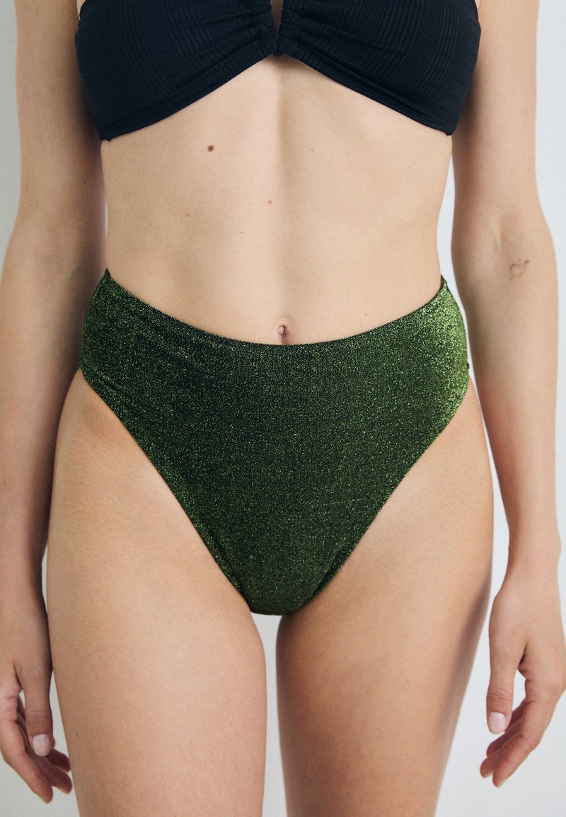 Aukštu juosmeniu bikini kelnaitės iš blizgaus tamsiai žalio audinio. Yra lygaus paviršiaus ir aukšto įpjovimo dizaino, kuris išryškina siluetą.