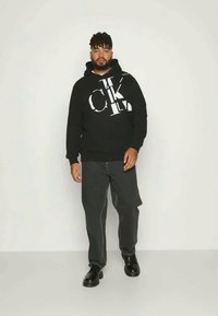 Sweatshirt preto com marca branca, calças largas cinzentas e sapatos pretos. A sweatshirt apresenta um cordão e punhos canelados, oferecendo um ajuste descontraído.