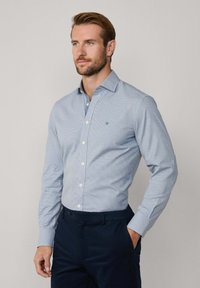 Muž s bradou, oblečený v světle modré košili s knoflíky a tmavomodrých kalhotách, stojící s jednou rukou v kapse na jednobarevném pozadí.