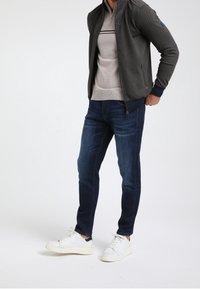 Giacca grigia con zip in tessuto testurizzato e dettagli blu navy, indossata sopra un maglione grigio chiaro; abbinata a jeans scuri e sneakers bianche.