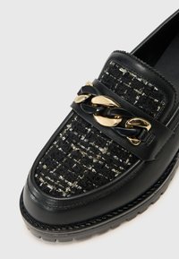 Musta loafers, joissa on tekstuurilla kudottu päällinen kultaisilla ja hopeisilla yksityiskohdilla. Paksu kultainen ja musta ketju koristaa remmiä. Kumiulkopohja.