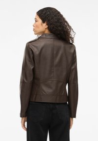 Hnedá kožená bunda s priliehavým strihom, obsahujúca límec, prešité detaily a zips na rukávoch, skombinovaná s čiernymi džínsami.