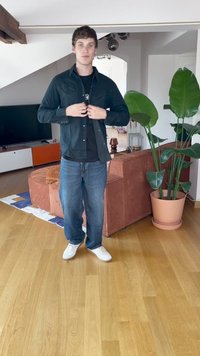 Juoda švarka už juodo marškinėlio, derinama su mėlynomis džinsomis ir baltomis sportbačiais. Fone yra augalas ir oranžinė sofa.