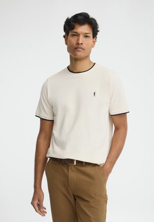 Hombre vistiendo una camisa beige de manga corta con ribete negro y pantalones marrones, de pie con una mano en el bolsillo contra un fondo liso.