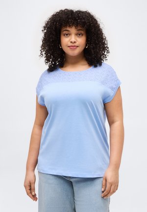 Kvinde med krøllet hår iført en lyseblå kortærmet top med hulmønster over brystet, kombineret med lyseblå jeans.