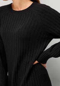 Pull noir en maille côtelée avec un col rond et des manches longues, présentant une surface texturée et une coupe ample. Orné de fentes latérales.