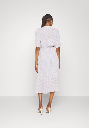 Femme aux cheveux tressés portant une robe midi fleurie violet clair et des sandales blanches à talons, debout de dos sur un fond uni.