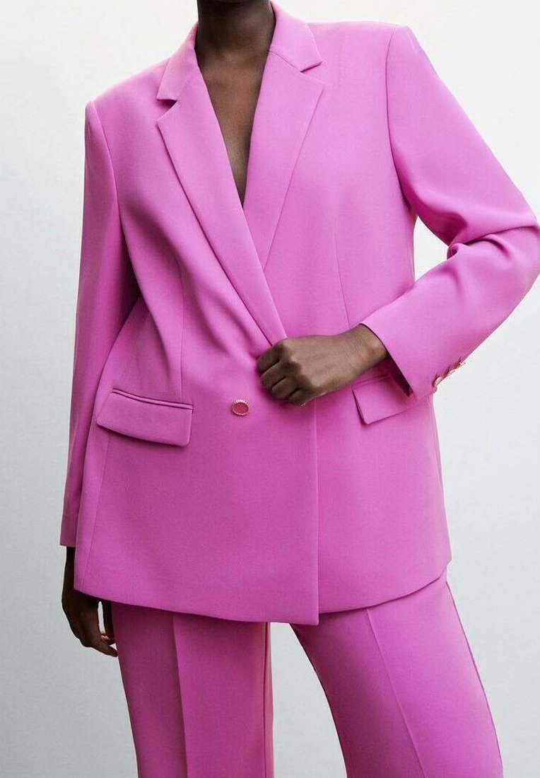Femme portant un tailleur rose vif sur mesure avec un blazer croisé et un pantalon droit assorti sur un fond uni.