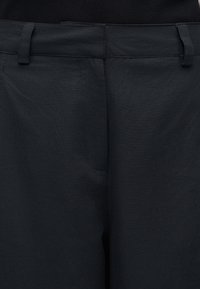 Κοντινή λήψη στη μέση σκούρου μπλε παντελονιού με θηλιές για ζώνη και εμπρόσθιο φερμουάρ, φορεμένο με μαύρο πουκάμισο κρυμμένο μέσα.