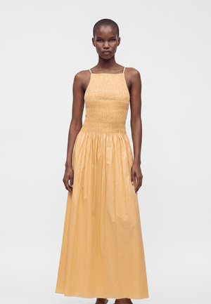 Femme aux cheveux courts portant une robe longue sans manches orange clair avec un corsage froncé et une jupe fluide, debout devant un fond uni.