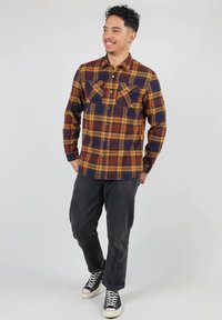 Chemise à manches longues à carreaux jaune, marine et bordeaux, avec poches poitrine et col boutonné. Portée avec un pantalon sombre et des baskets.