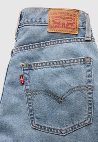 Πίσω τσέπη τζιν από ανοιχτό μπλε denim με πορτοκαλί ραφές, κόκκινη ετικέτα Levi's και ταμπέλα από δέρμα σε ταμπά χρώμα με τη μάρκα Levi Strauss & Co. πάνω από την τσέπη.