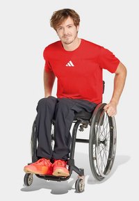 Röd sport-t-shirt med vit logotyp, svarta byxor och knallröda sneakers, bärs av en man i rullstol. Fokus på sportig design och passform.