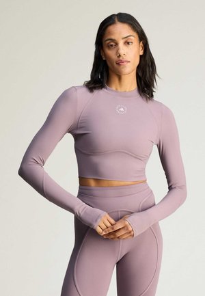 Kobieta o średnim odcieniu skóry, ubrana w długi, fioletowy top odsłaniający brzuch i legginsy z wysokim stanem, stojąca z rękami złożonymi przed sobą.