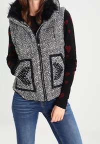 Sort-hvid tekstureret vest med pelsforet hætte, lynlås foran og mønstrede lommer. Båret over en sort sweater med røde hjerter.