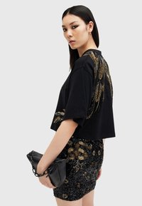 Svart cropped t-skjorte med gull- og sølvfjerdekorasjoner på skulderen, kombinert med et paljettbelagt svart skjørt og en svart clutch.
