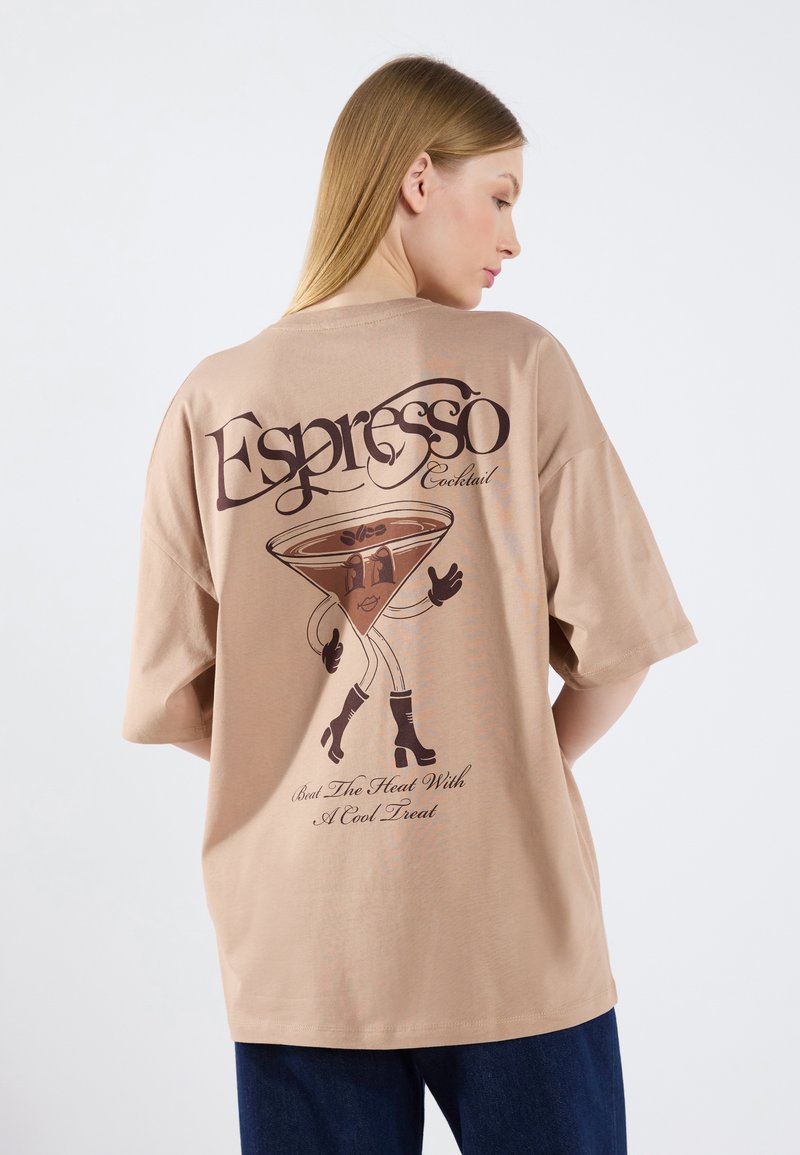 Camiseta de gran tamaño color beige con un gráfico de una copa de martini con piernas y brazos, junto a texto en marrón oscuro. Tejido de algodón, corte relajado.
