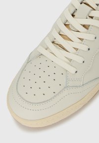 Close-up of beige sneaker toe with perforated leather, wide flat laces, and textured rubber sole on white background.