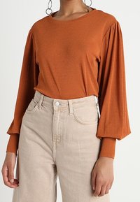 Rostfärgad långärmad topp med klockärmar, tillverkad av mjukt tyg. Kombinerad med högmidjade beige jeans med framfickor och subtila blekningar.