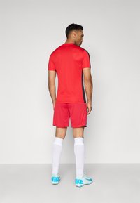 Punane spordijerseys lühikeste varrukatega, koos punaste lühikestega. Omab musti külgaktsente ja valgeid sokke, millele lisavad ilmet helesinised jalgpalliklubid.