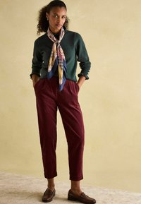 Dunkelgrüner Strickpullover, kastanienbraune Cordhose, gemusterter Schal, braune Slipper; mit Knopfdetails an den Ärmeln.