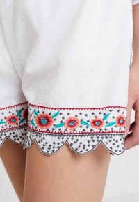 Bílé šortky s texturovanou látkou, s vroubkovaným lemem a vyšívaným květinovým motivem v červené, tyrkysové a černé barvě poblíž stehna.