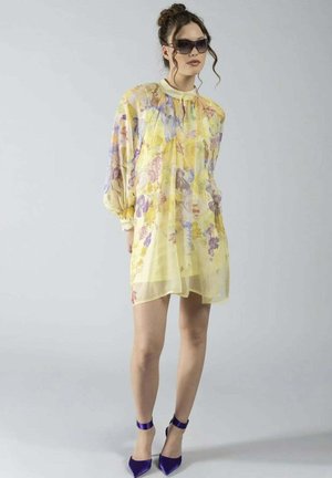 Donna che indossa un abito giallo a fiori trasparente, tacchi viola con cinturino alla caviglia e grandi occhiali da sole scuri, in piedi contro uno sfondo chiaro e uniforme.