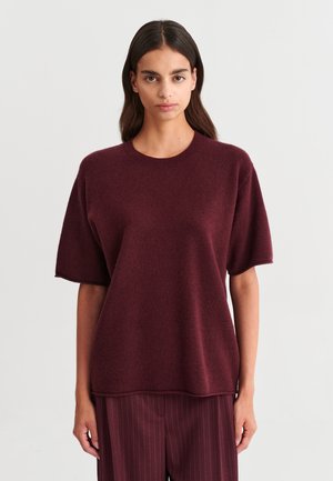 Jeune femme aux longs cheveux foncés portant un pull ample à manches courtes bordeaux et un pantalon bordeaux à fines rayures, debout devant un fond uni.