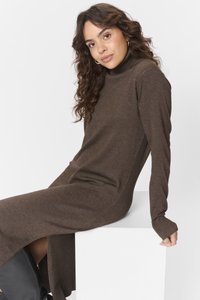 Robe turtleneck marron en tricot doux, dotée de longues manches et d fentes latérales, posée sur un bloc blanc.