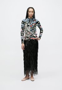 Pull à manches longues avec un motif multicolore de tigre et d'étoiles ; associé à une jupe longue noire à franges ; chaussures plates de couleur claire.