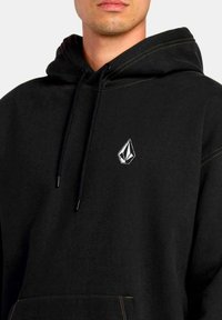 Sudadera negra con bolsillo tipo canguro, con un logo blanco en el lado izquierdo del pecho y costuras amarillas en contraste en la capucha.